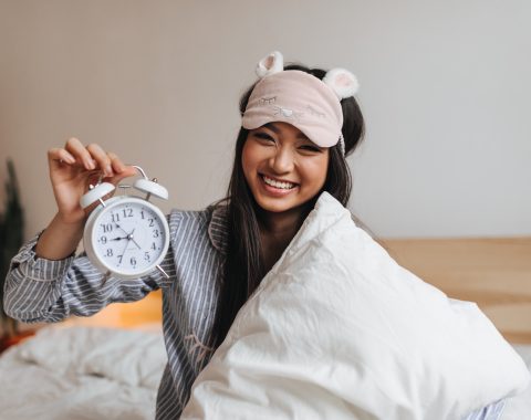 Smiling woman holding an alarm clock in bed, wearing a sleep mask, representing the importance of sleep duration and regularity for better sleep health